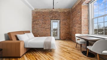 A modern bedroom with a brick wall, a wooden floor, a bed, a sofa, a table, and a chair.