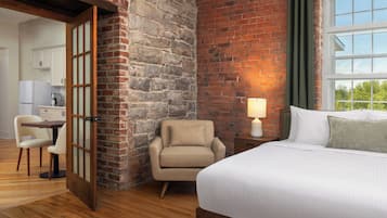 A hotel room with a brick wall, a bed with white linens, a chair, a small table with a lamp, and a view through a window.