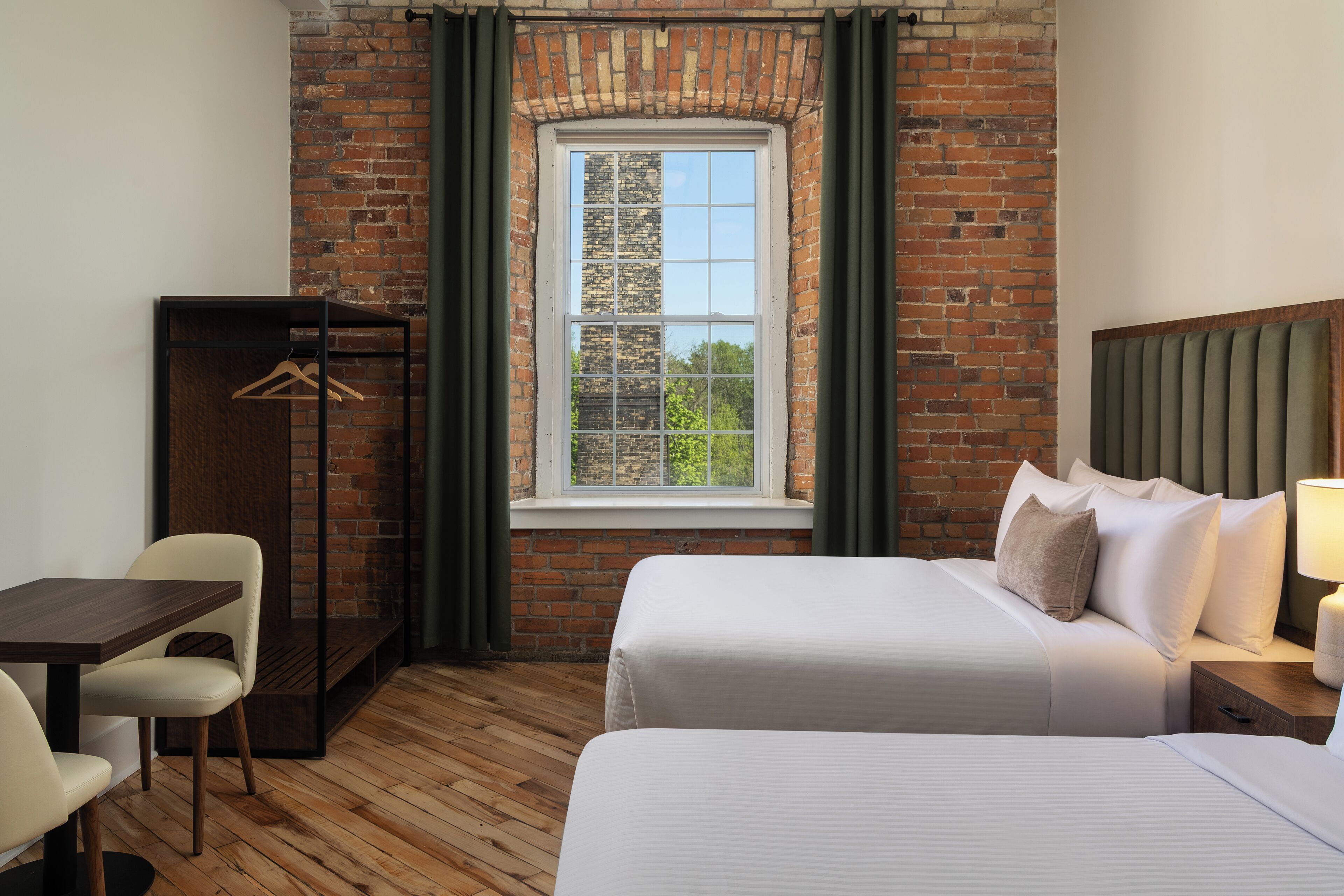 A hotel room with two beds, a desk, a wardrobe, and a window with a view of trees.