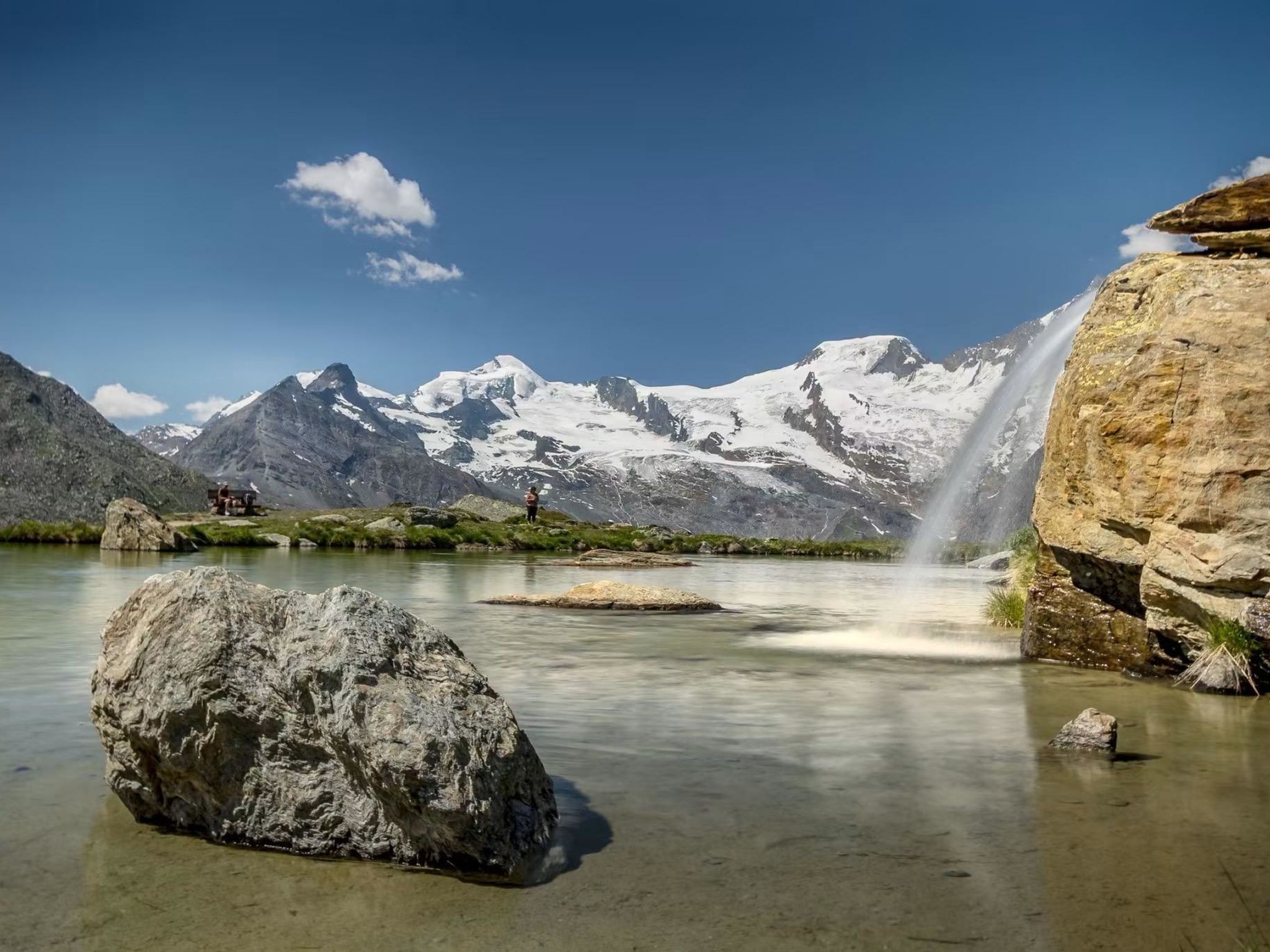 Paisaje montañoso con cielo despejado, cascada y un lago rocoso.
