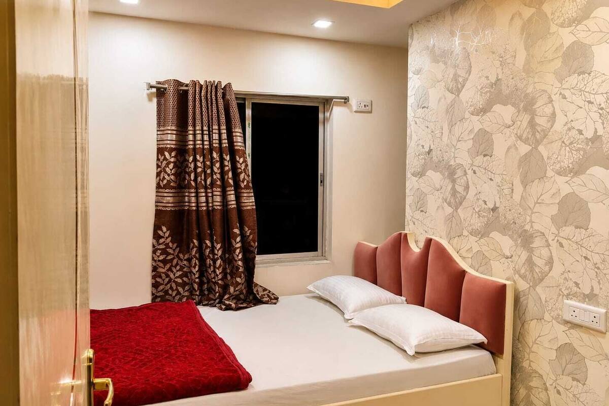 A single bed with a red blanket, a window with a curtain, and a wall with a leaf pattern.