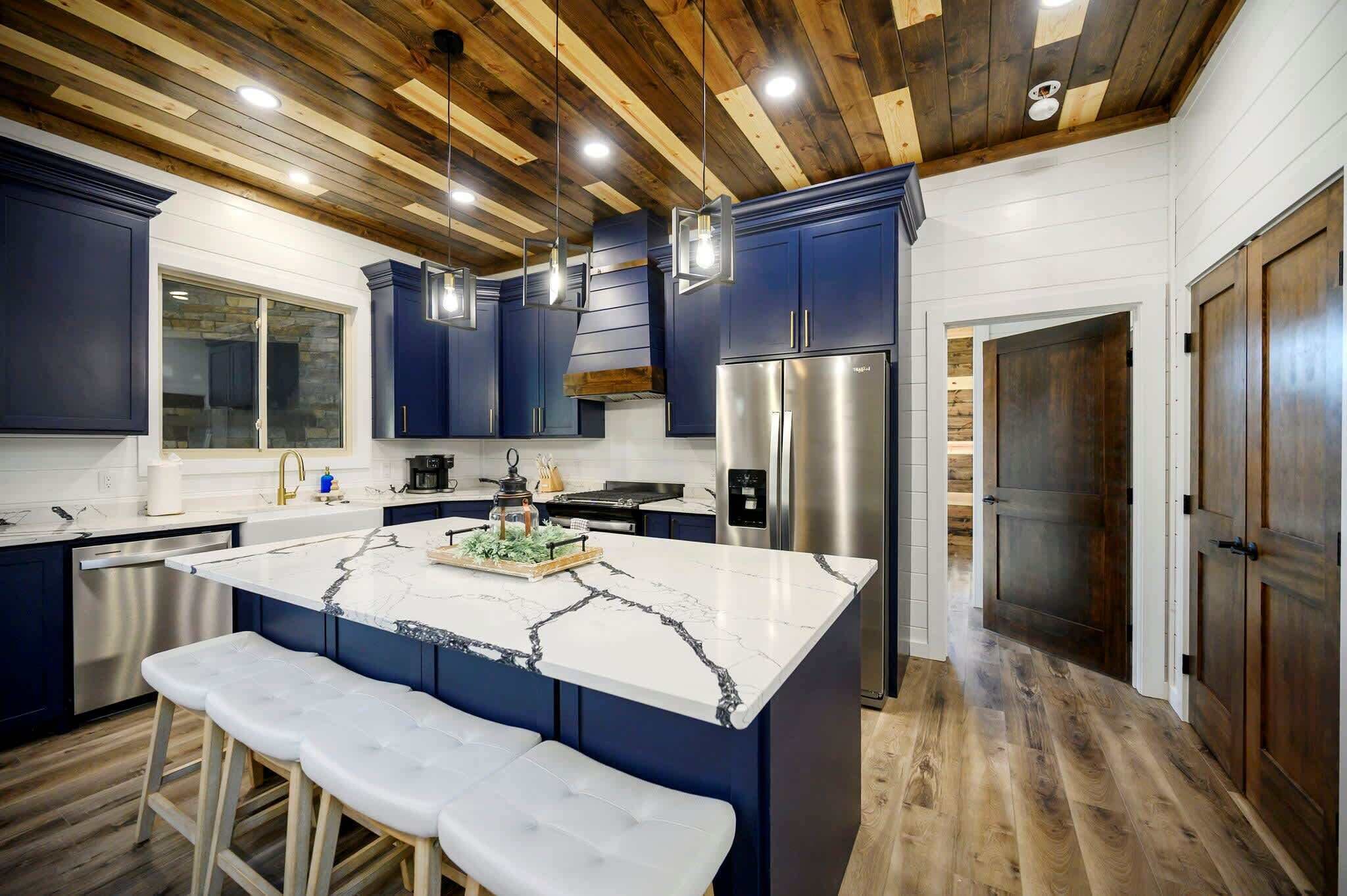 A modern kitchen with dark blue cabinets, a marble island, stainless steel appliances, and wooden ceiling beams.