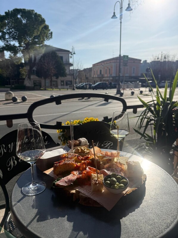 A table with a variety of food items and two glasses of wine, set outdoors with a view of a street and buildings in the background.