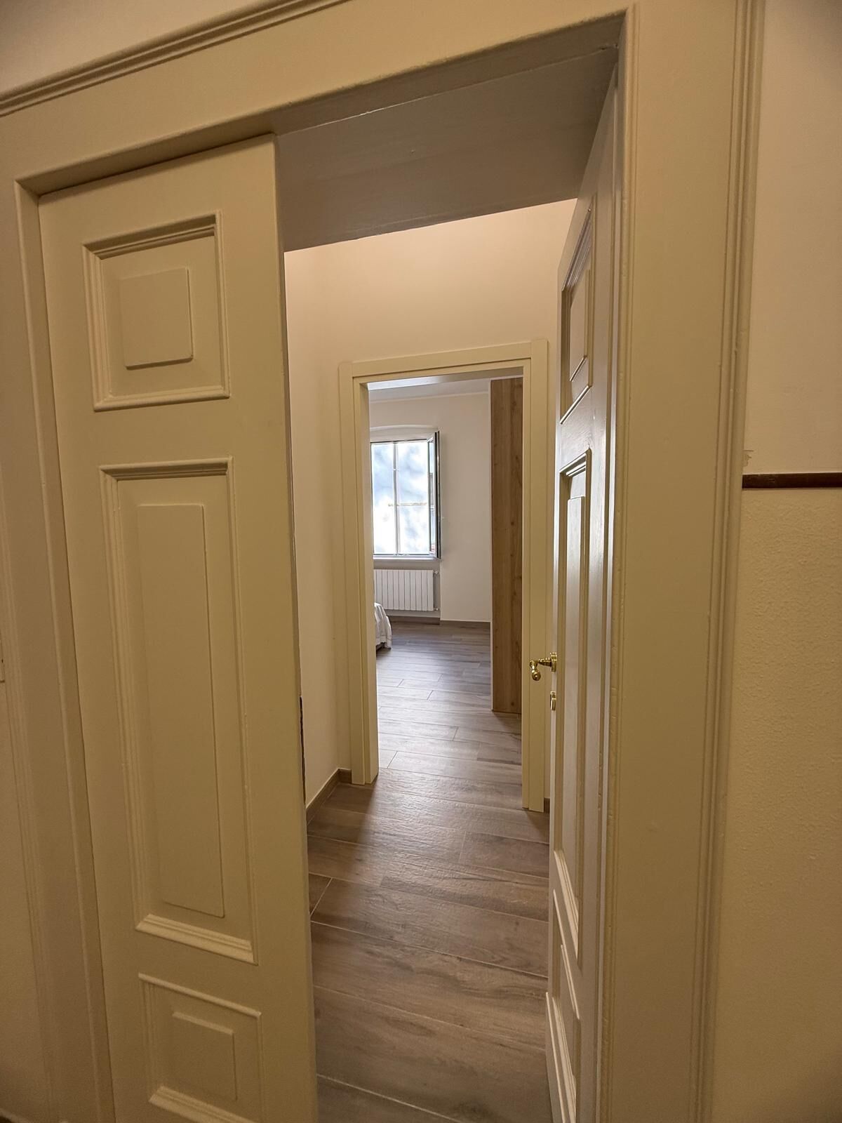 A hallway with wooden flooring and cream-colored walls, leading to a room with a large window and a radiator.
