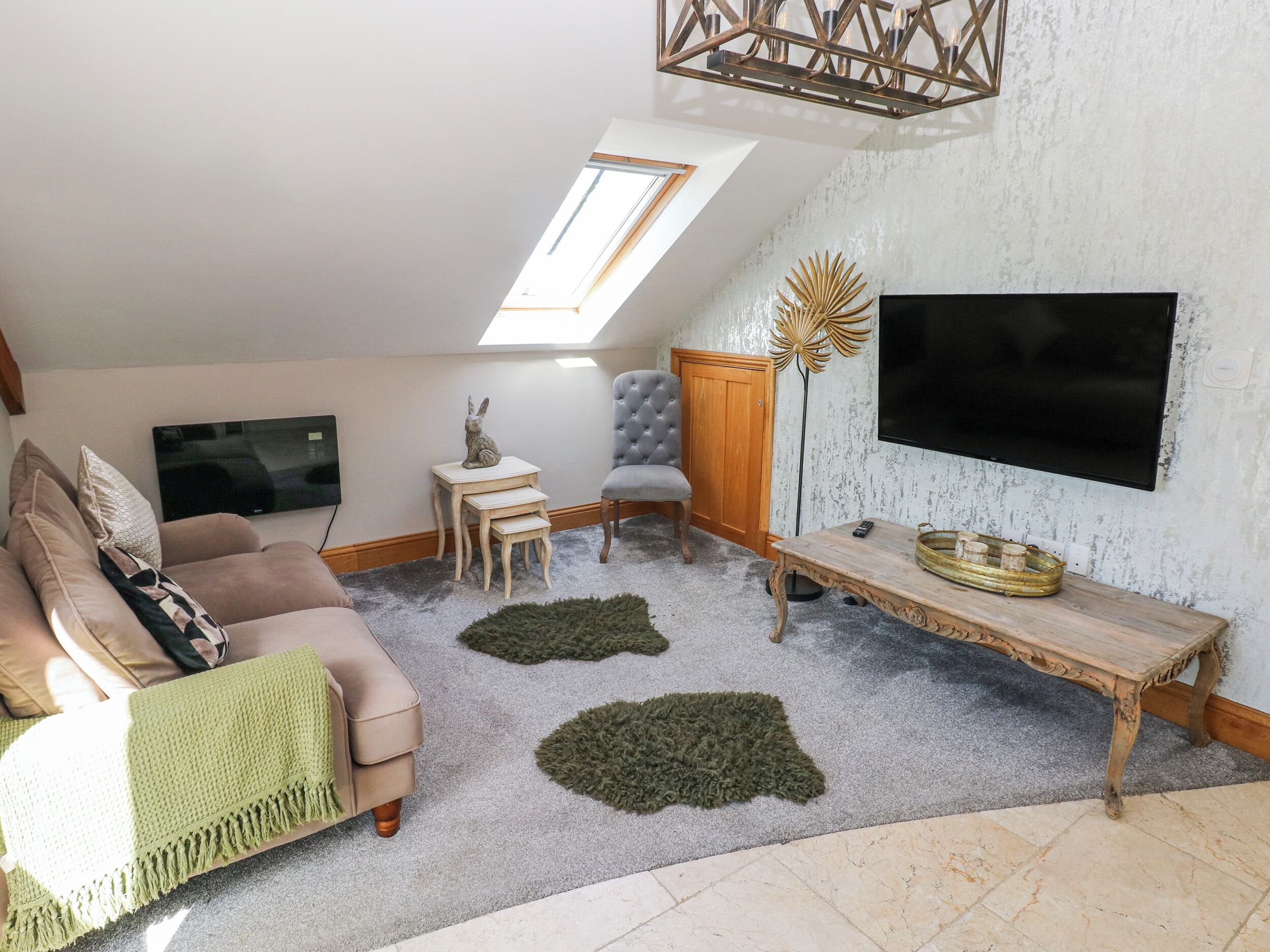 A loft-style living room with a high ceiling, a skylight, a flat-screen TV mounted on the wall, a sofa, a chair, a small table, and a decorative floor rug.