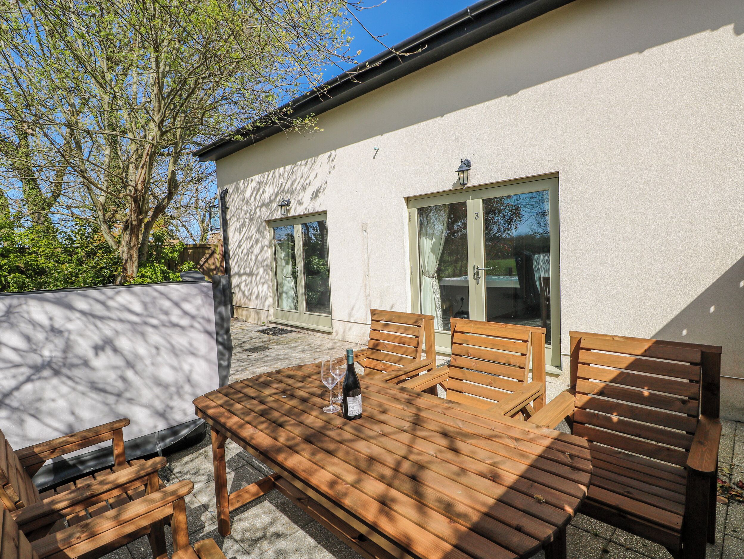 A wooden patio with a table and benches, a sliding glass door, and a white exterior wall.