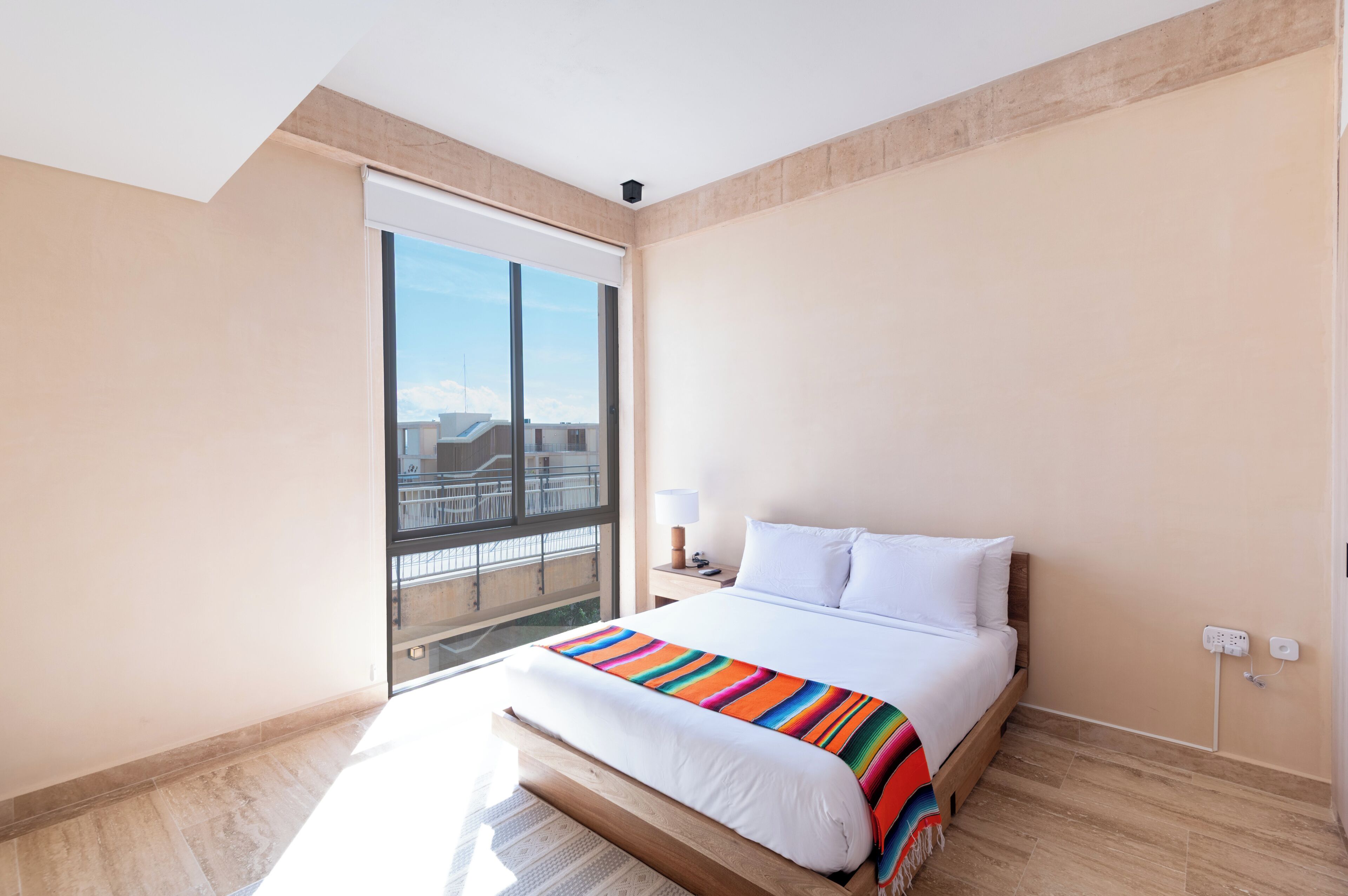 A hotel room with a large bed, a nightstand, a lamp, a window with a view of buildings, and a power outlet.