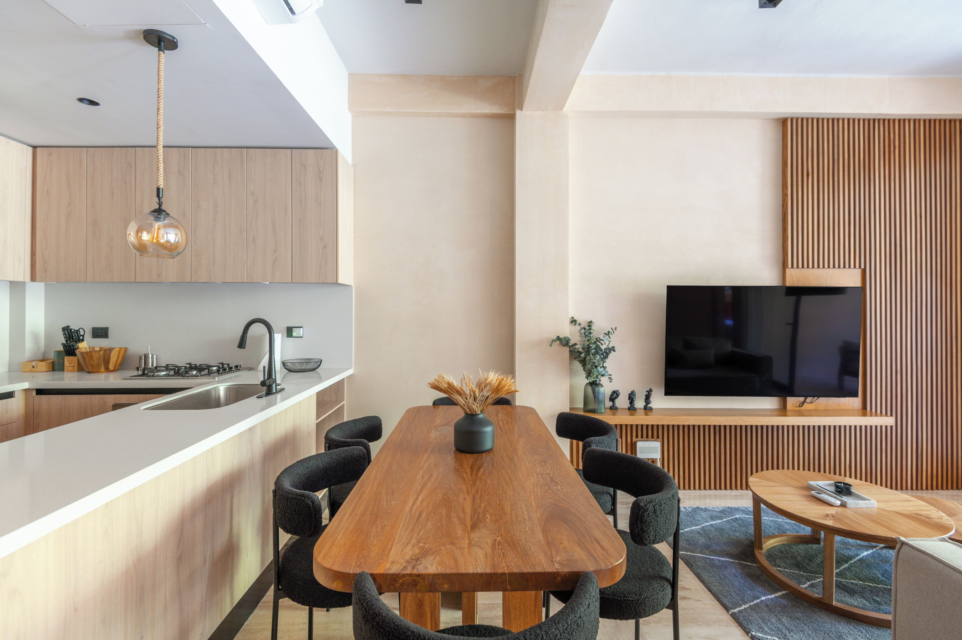 A modern kitchen with a central island, a dining area with a wooden table and chairs, and a flat-screen TV mounted on a wooden panel wall.