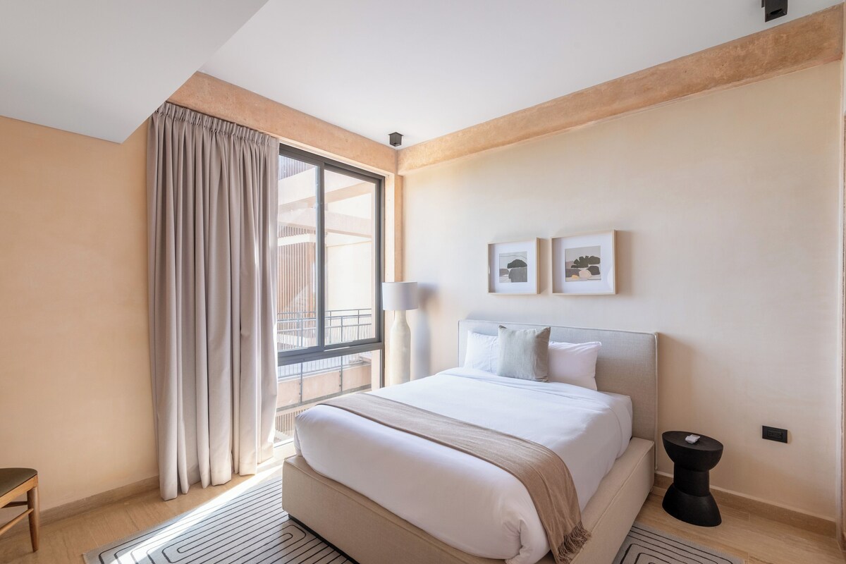 A hotel room with a bed, a window with curtains, a bedside table, and a wall-mounted picture.