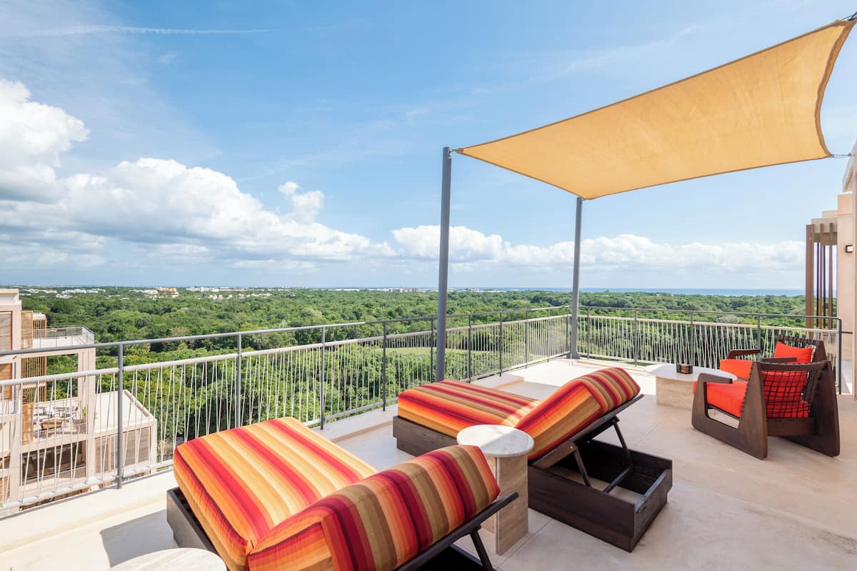 A balcony with two lounge chairs, a table, and a large canopy. The balcony overlooks a lush green landscape.