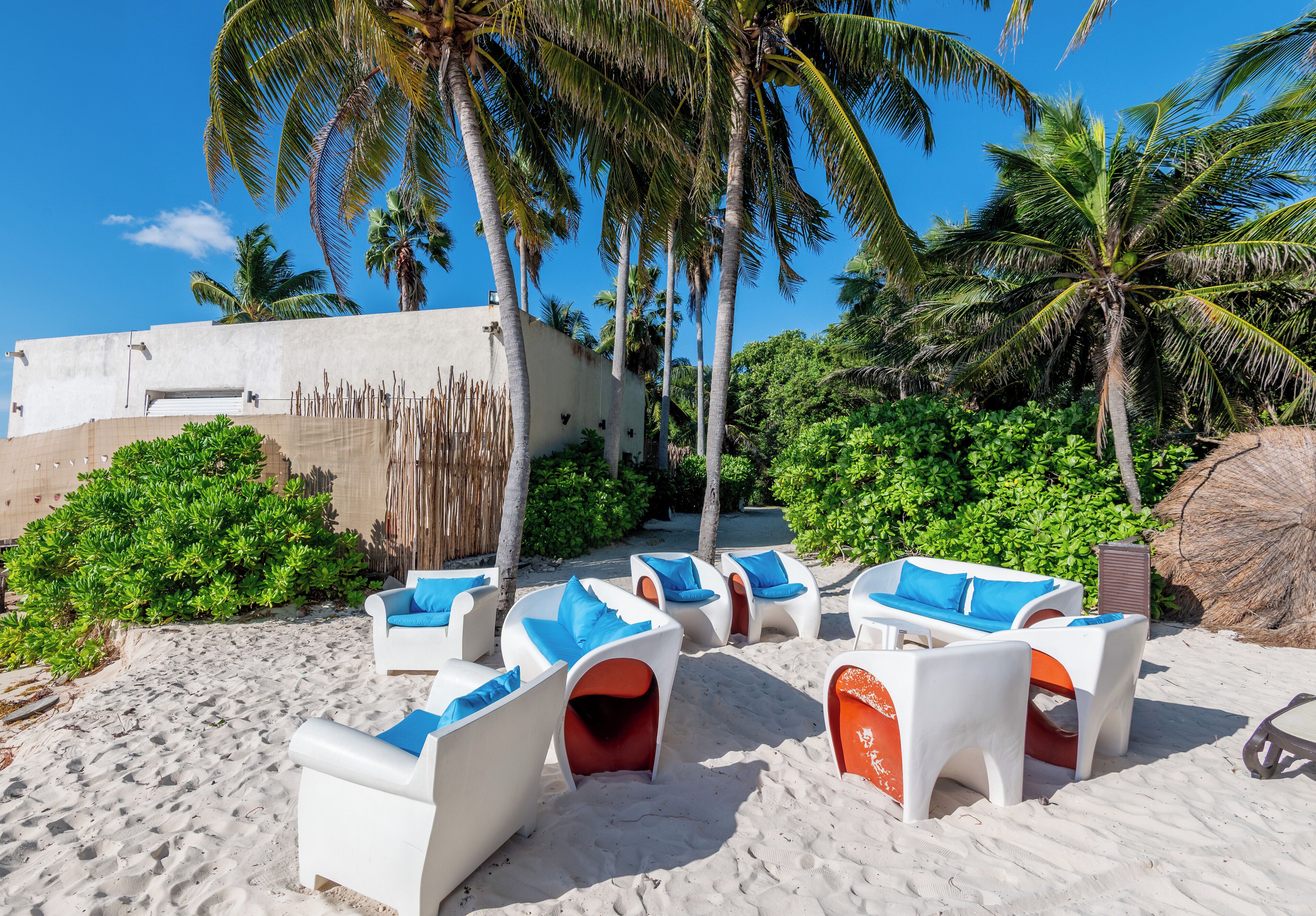 Un espace de détente aménagé au bord de la plage, avec des chaises longues blanches et des coussins bleus, entouré de palmiers et bordé d’un bâtiment en béton.