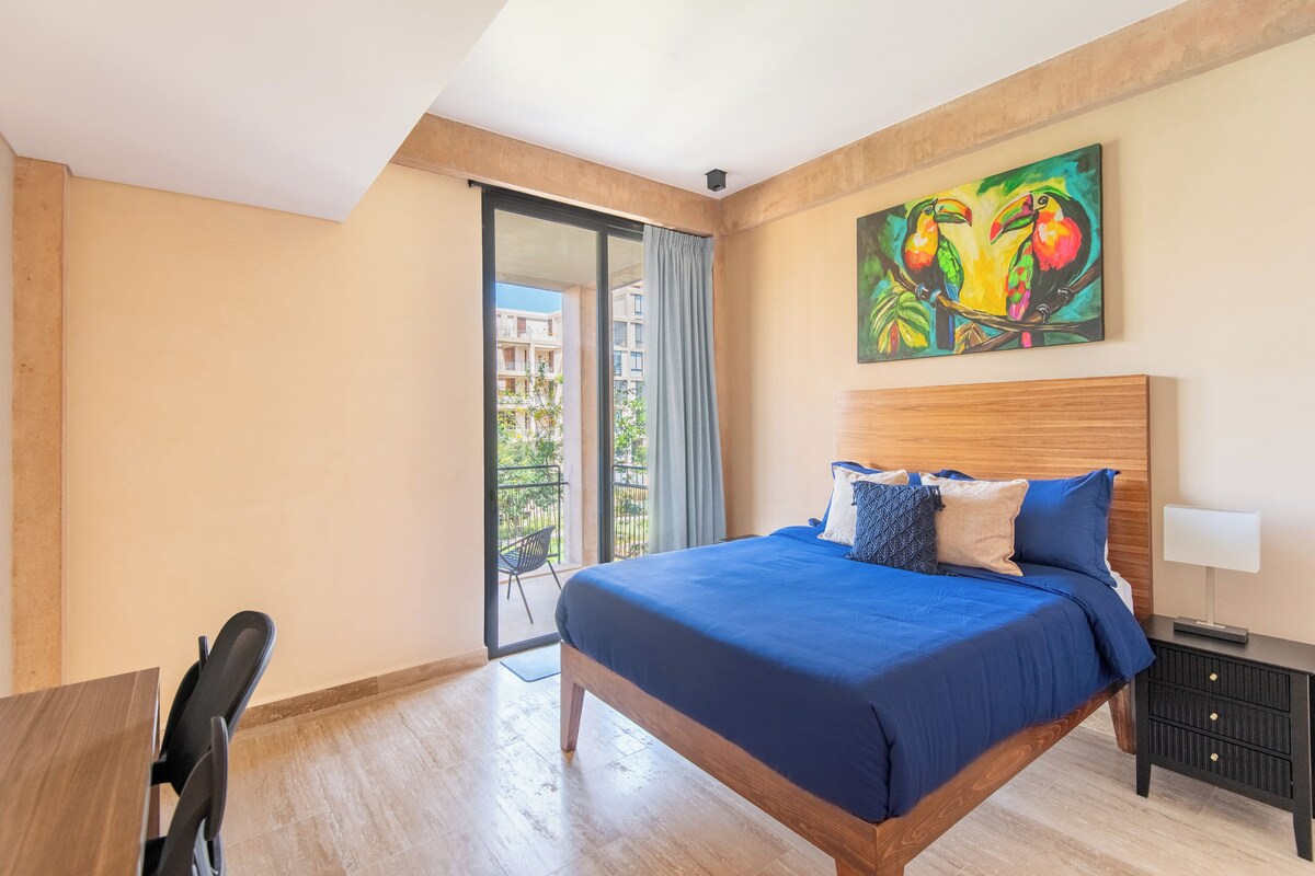 A hotel room with a large bed, a desk, and a chair. There is a balcony with a view of buildings.