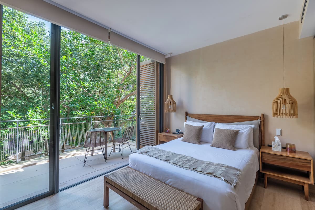 A modern bedroom with a large bed, a wooden nightstand, a bedside table, and a view of a balcony with a table and chairs.