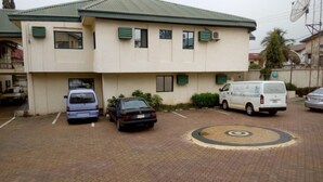 A building with a flat roof, multiple windows, and a paved parking area with several vehicles. - DAVIES SUITES  (Enugu)