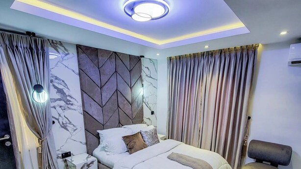 A modern bedroom with a bed, curtains, a nightstand, and a chair. - DAVIES SUITES  (Enugu)