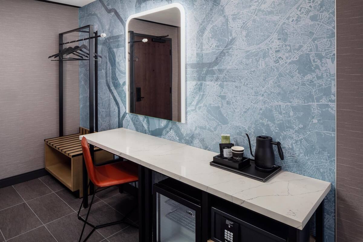 Een compacte hotelkamer met een lang wit aanrecht, een kleine koelkast, een zithoek met een rode stoel, een handdoekhouder en een spiegel met houten deur.