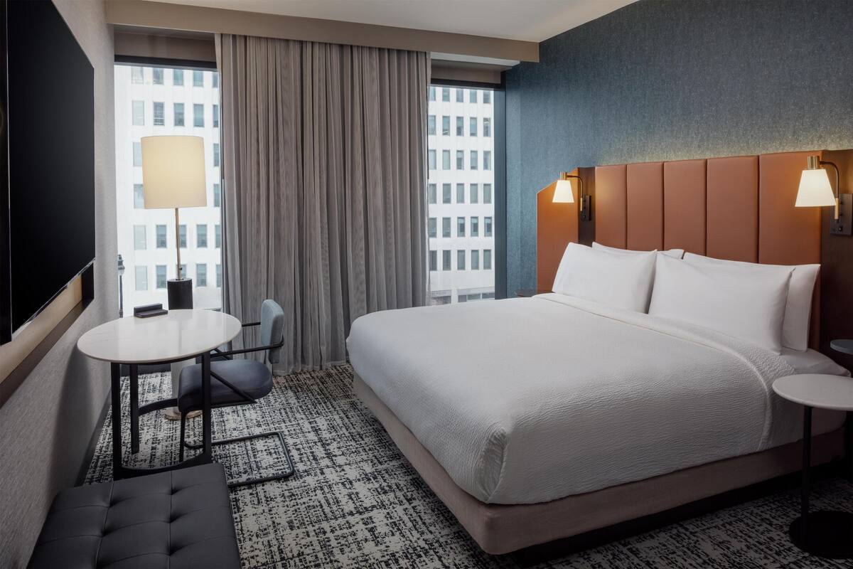 Hotelkamer met een groot bed, een klein rond tafeltje, een stoel, een bank, een raam met gordijnen en uitzicht over de stad.