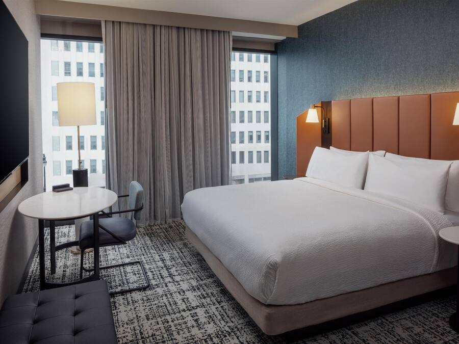 Hotelkamer met een groot bed, een klein rond tafeltje, een stoel, een bank, een raam met gordijnen en uitzicht over de stad.