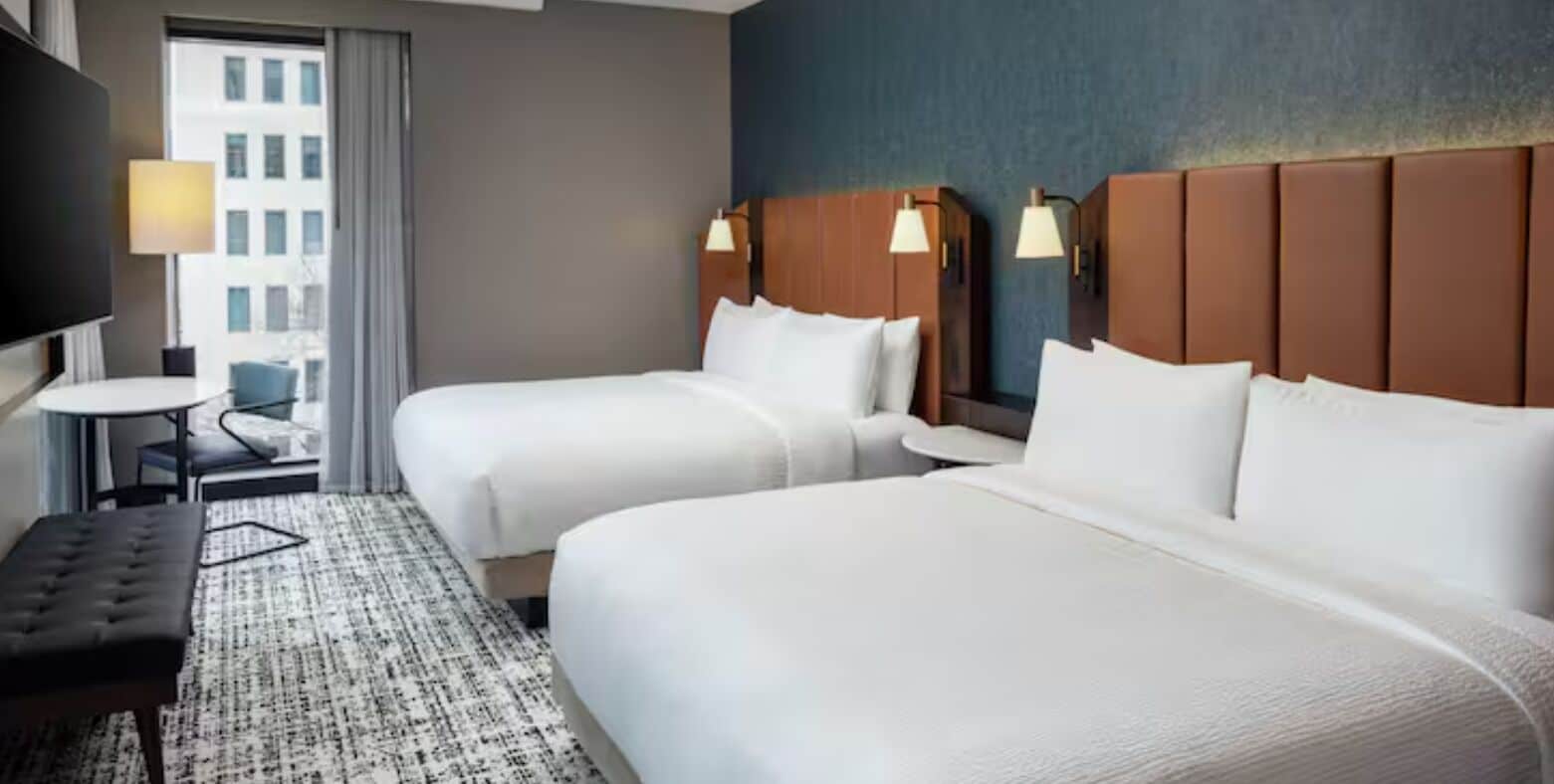 A hotel room with two beds, a desk, a chair, and a TV.