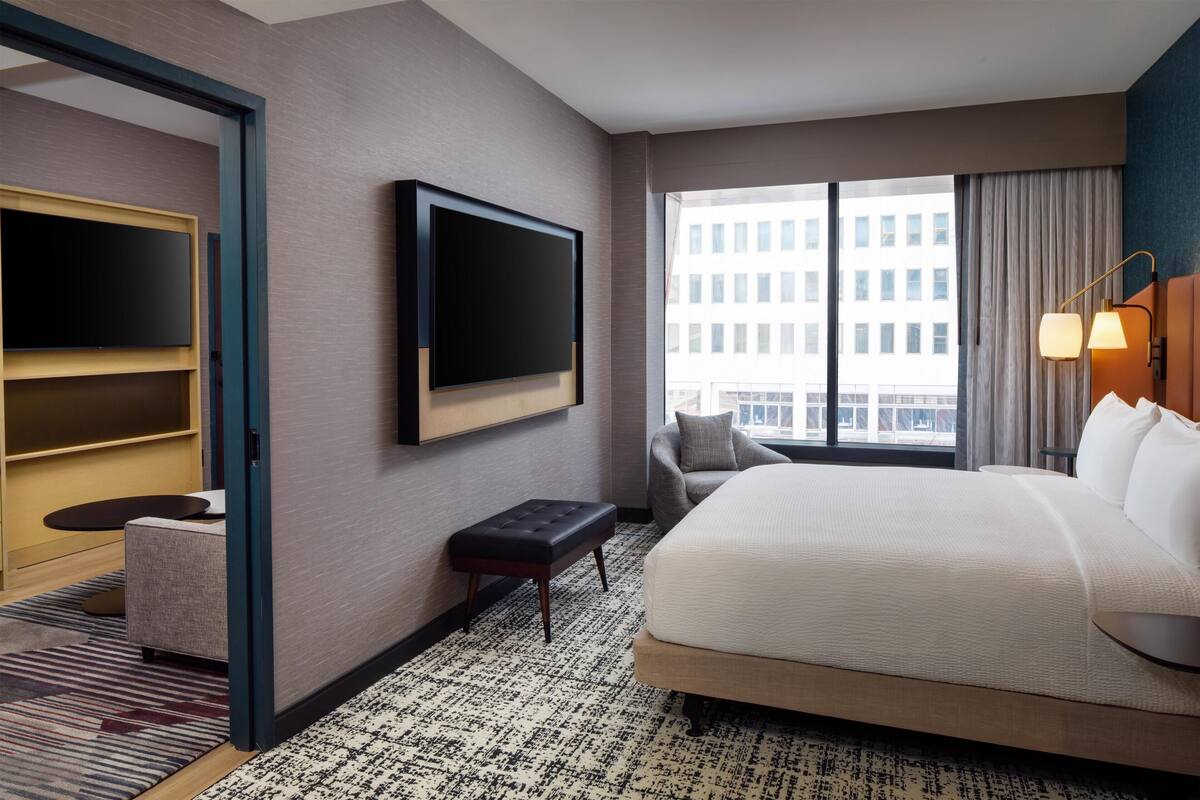 Hotelkamer met een bed, een aan de muur bevestigde televisie, een stoel, een klein tafeltje en een raam met uitzicht op gebouwen.