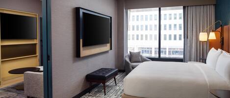 A hotel room with a bed, a TV mounted on the wall, a chair, a small table, and a window with a view of buildings.
