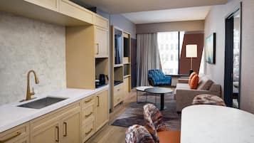 Quarto de hotel moderno com cozinha, área de estar com sofá e poltronas, uma mesinha e uma janela grande.