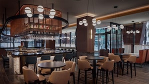 Zona de bar moderna con mesas y sillas redondas, un mostrador central y lámparas esféricas colgantes.