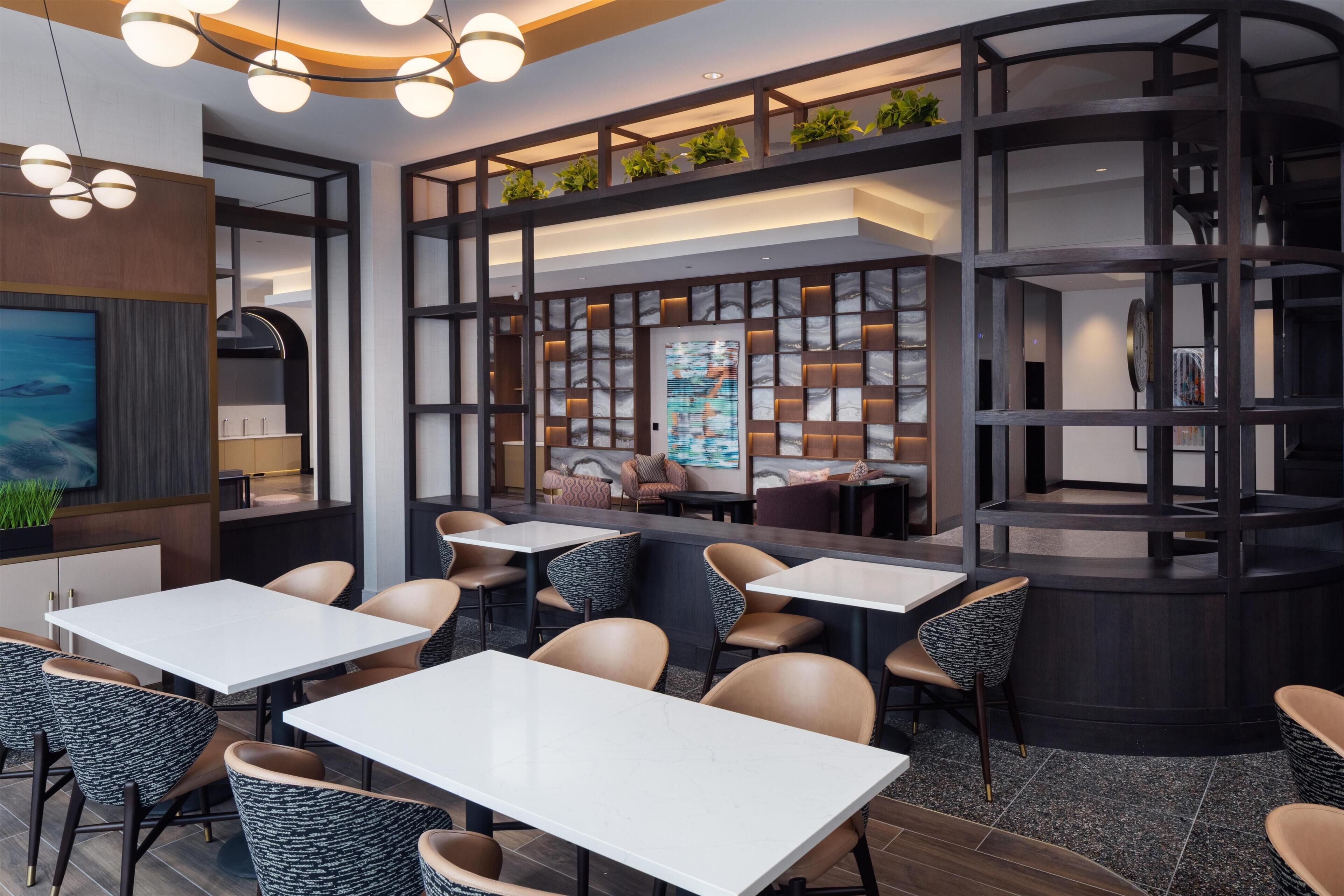 A modern restaurant interior with white tables, cushioned chairs, a central seating area with sofas, and a decorative wall with plants.