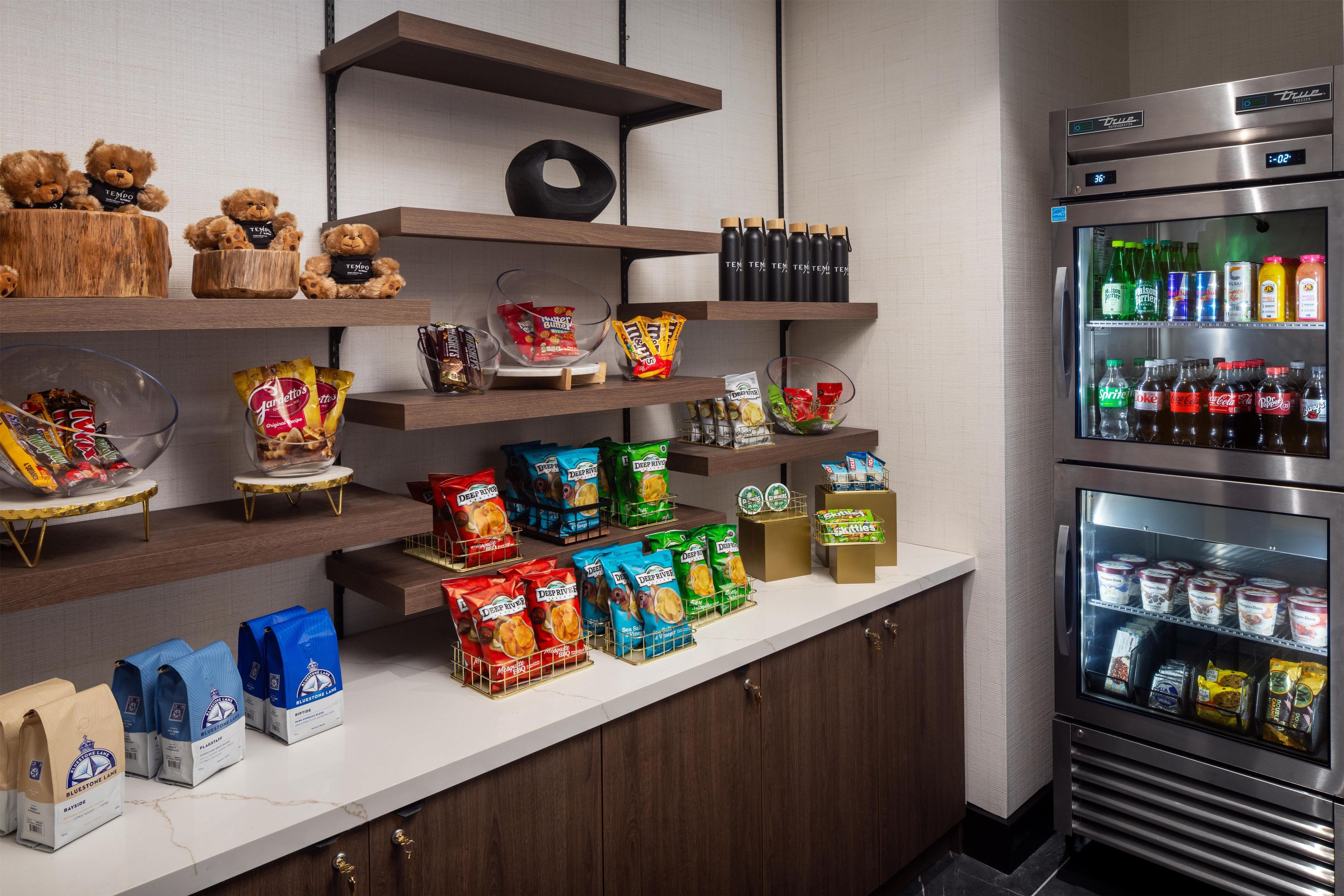 Una kitchenette con un refrigerador lleno de bebidas, una cafetera y diversos bocadillos y condimentos.