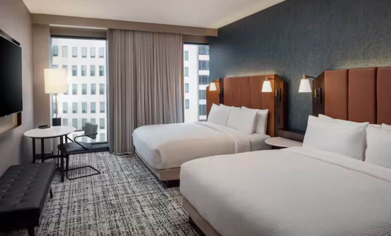 A hotel room with two beds, a desk, a chair, a TV, and a view of a building through the window.