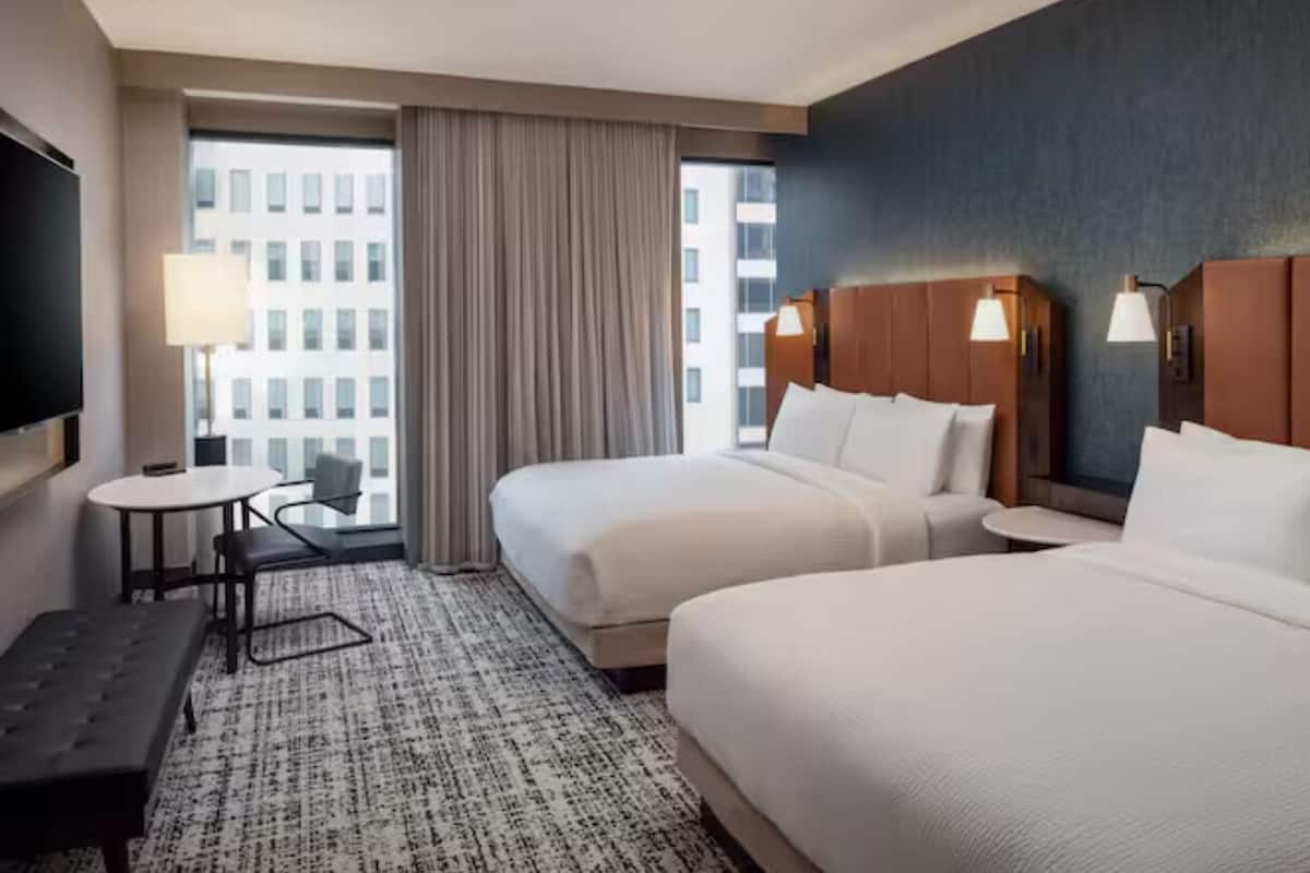 A hotel room with two beds, a desk, a chair, a TV, and a view of a building through the window.