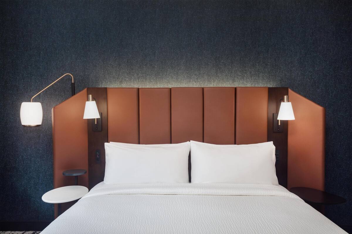 Een hotelkamer met een bed, twee bedlampen, een klein rond tafeltje en een bedieningspaneel aan de muur.