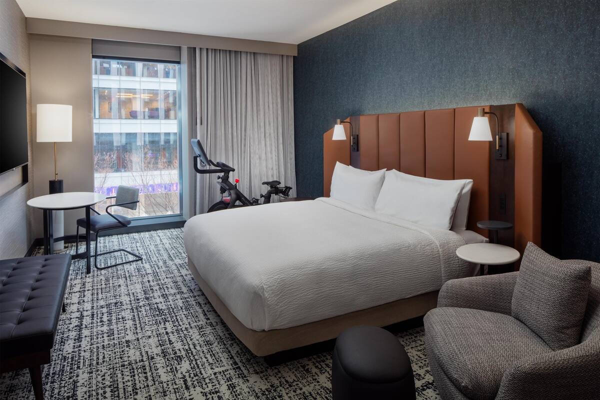 Hotelkamer met een groot bed, een zithoek met een fauteuil en poef, een tafeltje en een televisie.