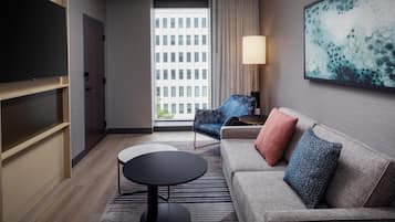 A modern hotel room with a sofa, a small round table, a TV, and a large window with a city view.