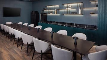 Sala de conferências com uma mesa comprida, cadeiras brancas, uma TV de tela plana e uma parede azul com prateleiras.