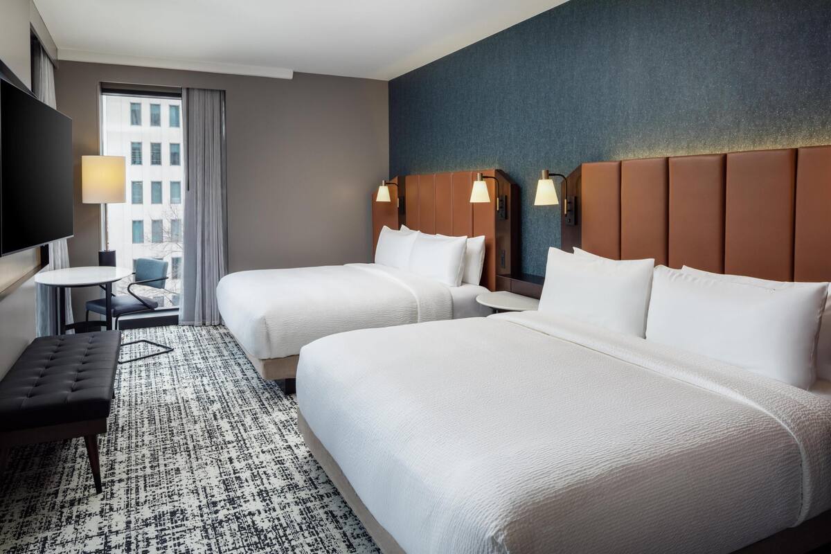 Een hotelkamer met twee bedden, een televisie, een klein tafeltje en uitzicht op een gebouw.