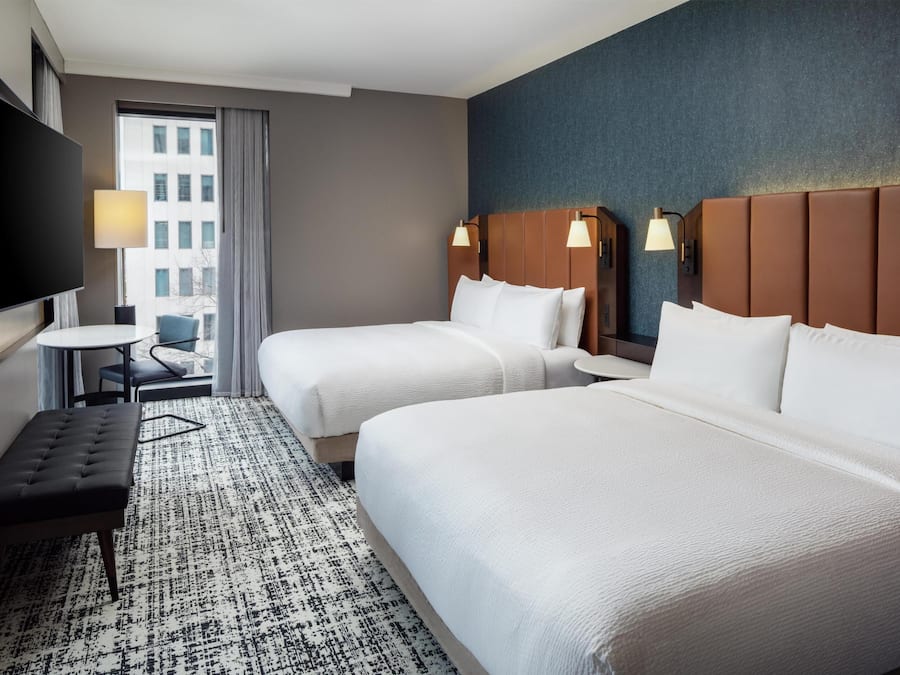 A hotel room with two beds, a TV, a small table, and a view of a building.