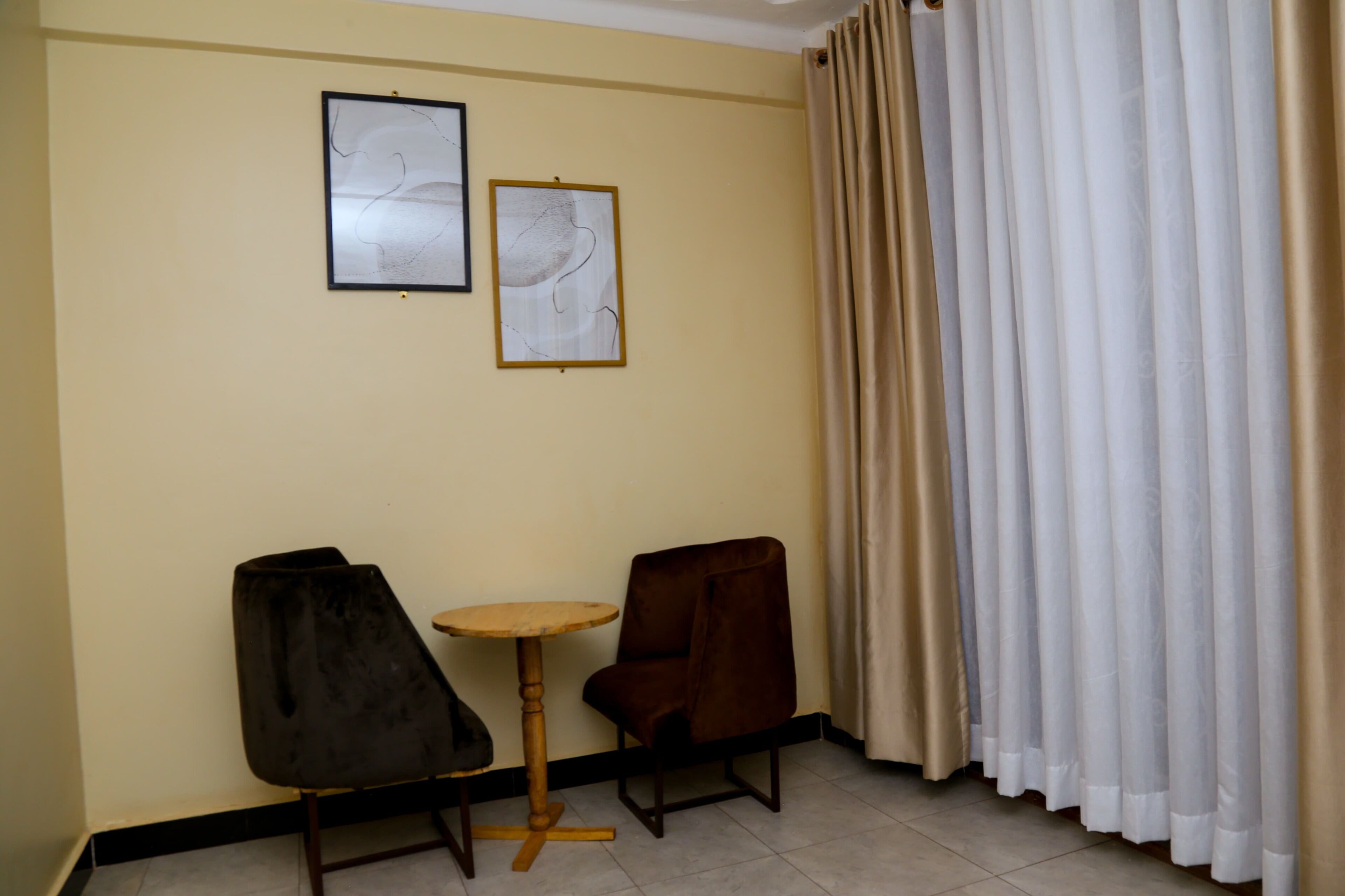 A room with two framed abstract artworks on the wall, a brown chair, a small round table, and a beige curtain.