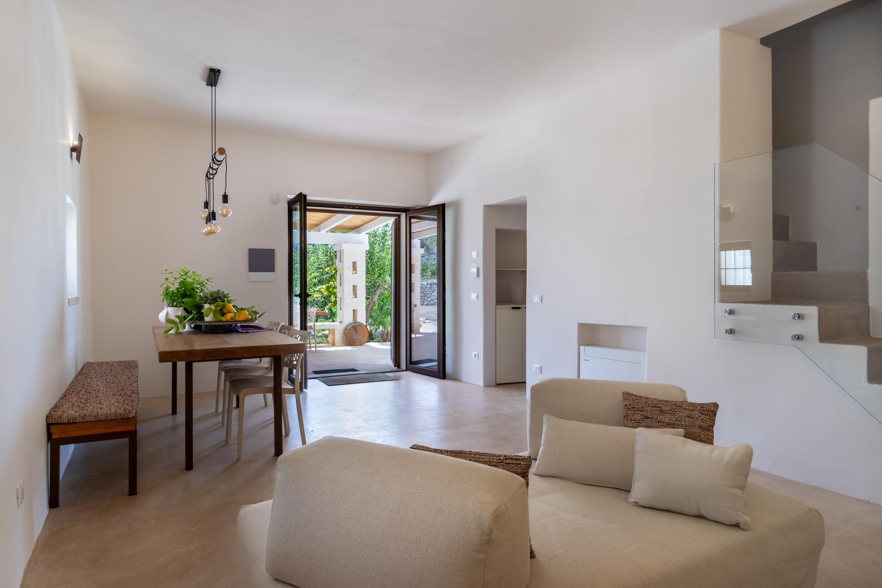 A modern living room with a sofa, a dining table with chairs, and a bench. There is a staircase to the right and a door leading to an outdoor area.