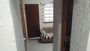 Un couloir menant à une pièce où se trouvent un canapé, une porte et une fenêtre avec des stores.