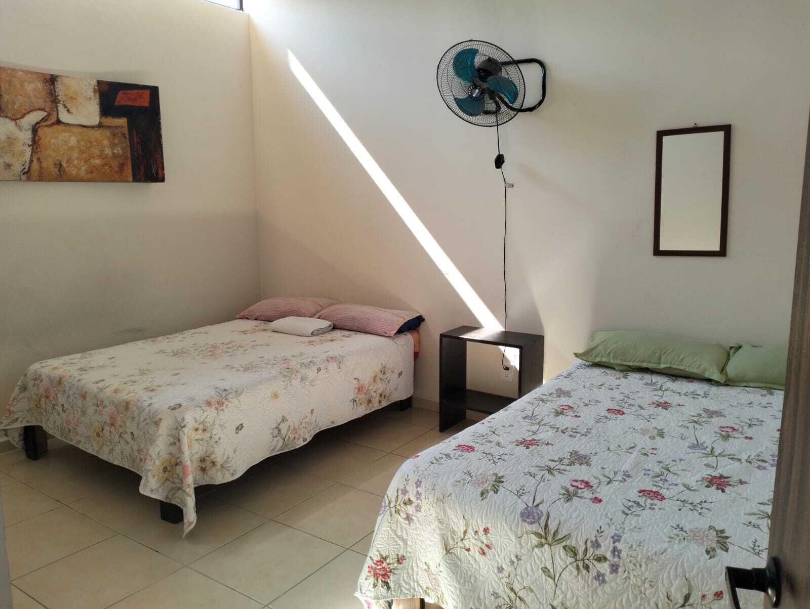 Ein kleiner, kompakter Raum mit zwei Einzelbetten, einem Standventilator, einem kleinen Tisch und einem gerahmten Bild an der Wand.