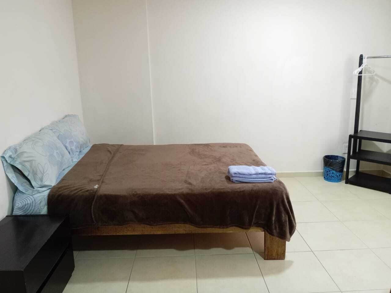 Una cama individual con cubrecolor café, almohada azul y una toalla doblada en el cabecero. Un mesita de noche negra está a la izquierda de la cama.