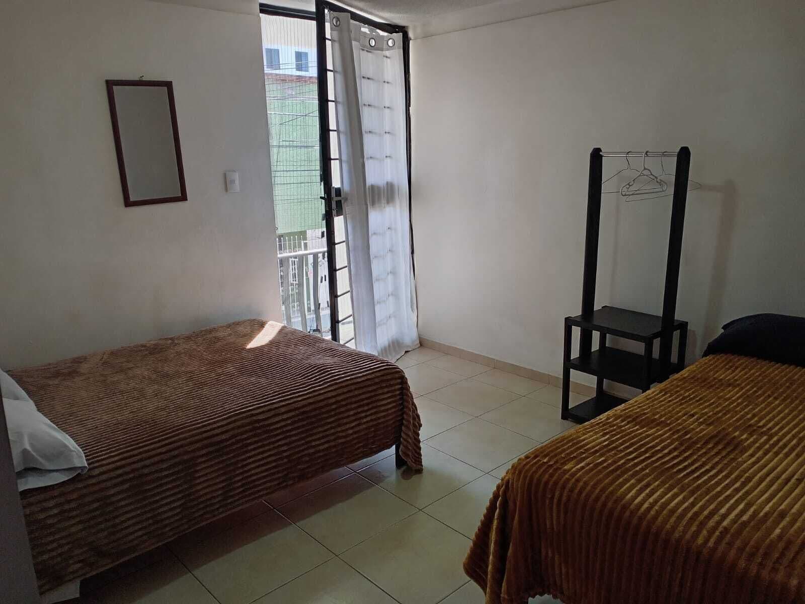 A small, clean room with two single beds, a chair, a hanging clothes rack, and a window with a view of a building outside.