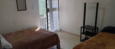 Una habitación pequeña y limpia con dos camas individuales, una silla, un perchero y una ventana con vistas a un edificio.