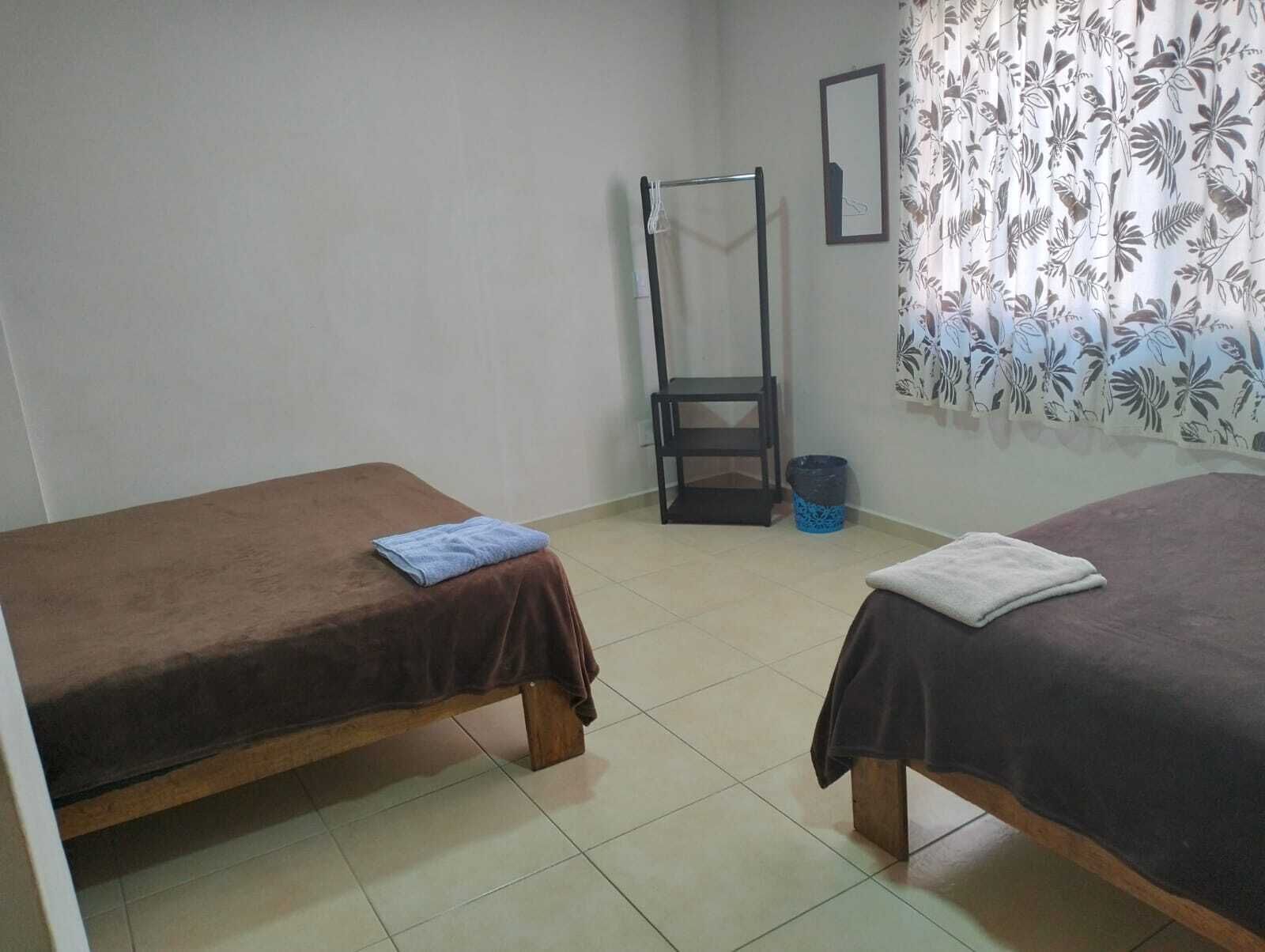 Una habitación con dos camas individuales, una mesita, una silla, un cubo de basura y una cortina con estampado de hojas.