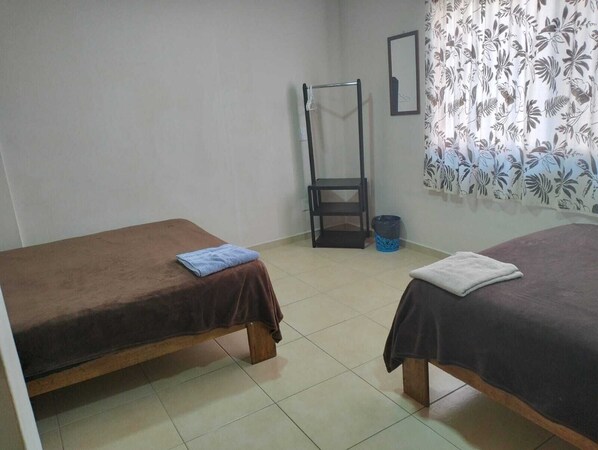 A room with two single beds, a small table, a chair, a trash bin, and a curtain with a leaf pattern. - Hotel Morelia Nova (Morelia)