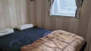 Una cama individual con un colchón azul oscuro y una colcha color beige. Hay dos toallas blancas enrolladas sobre la cama. La habitación tiene paredes con rayas verticales y una ventana con cortinas.