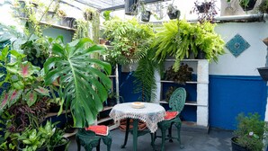 Un pequeño patio con una mesa redonda y dos sillas, rodeado de macetas con diferentes plantas y una pared azul y blanca.