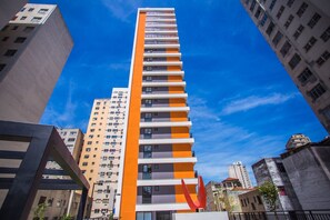 A tall, modern building with a distinctive orange and white color scheme, surrounded by other buildings. - Studio no All You Need - All0604 (Curitiba)