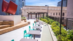 An outdoor seating area with tables and chairs, a red sculpture, and a building with classical architecture in the background. - Studio no All You Need - All0604 (Curitiba)